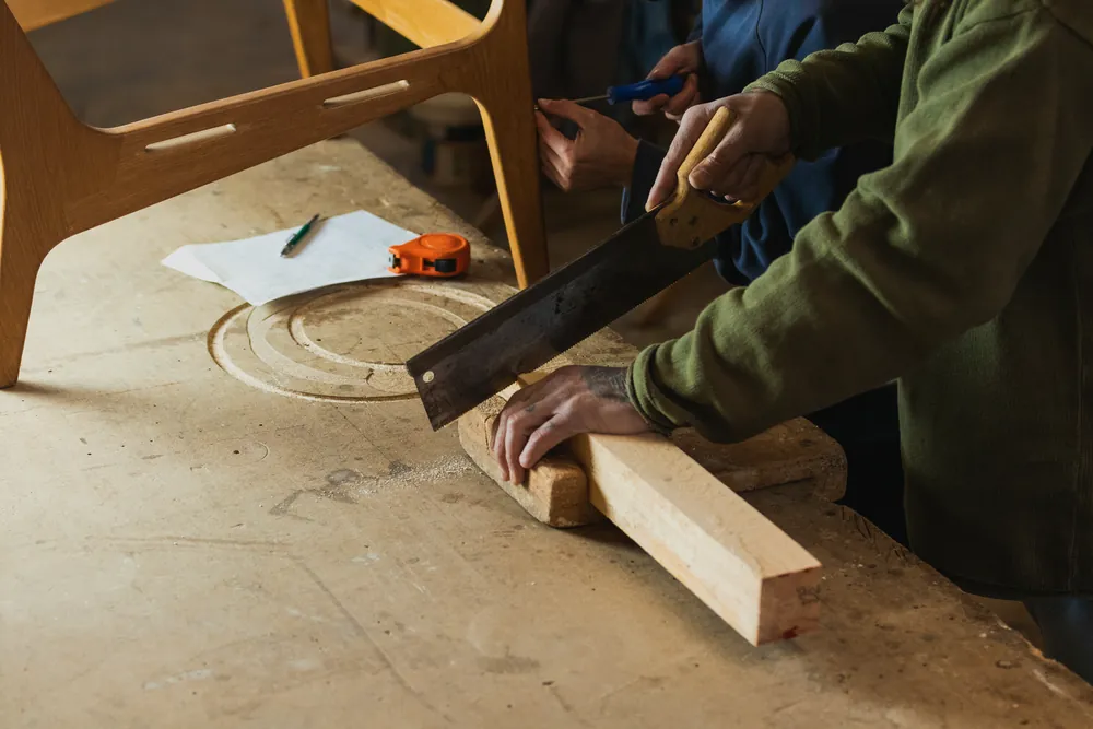 Two makers cutting a timber component at a workshop bench.